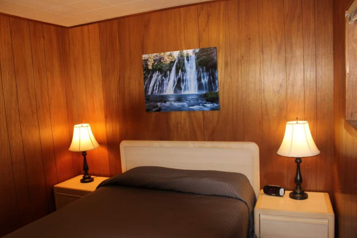 Habitación de hotel con una cama, dos lámparas de noche, un reloj digital y un cuadro colgado en la pared que representa una cascada.