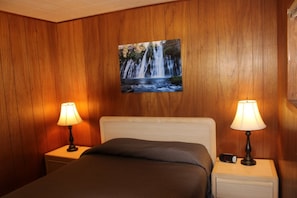 A hotel room with a bed, two bedside lamps, a digital clock, and a wall-mounted painting of a waterfall.