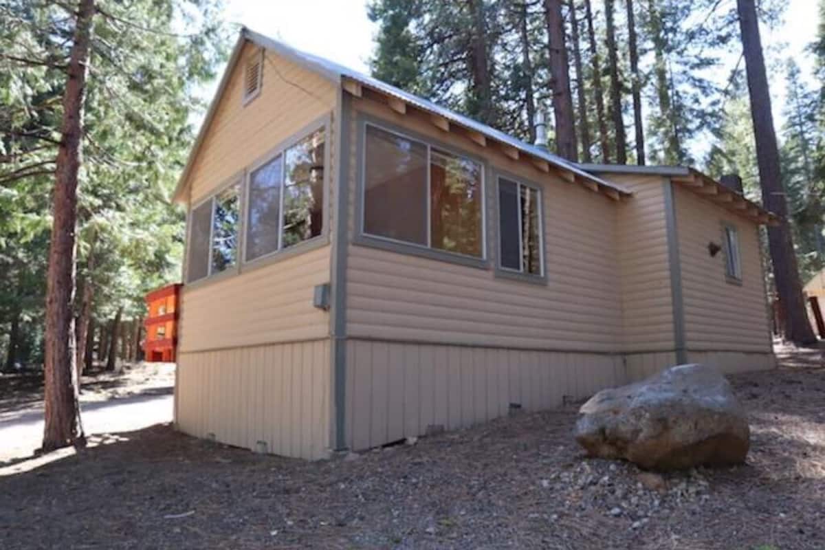 Una cabaña pequeña de un piso, con ventanas grandes, rodeada de árboles.