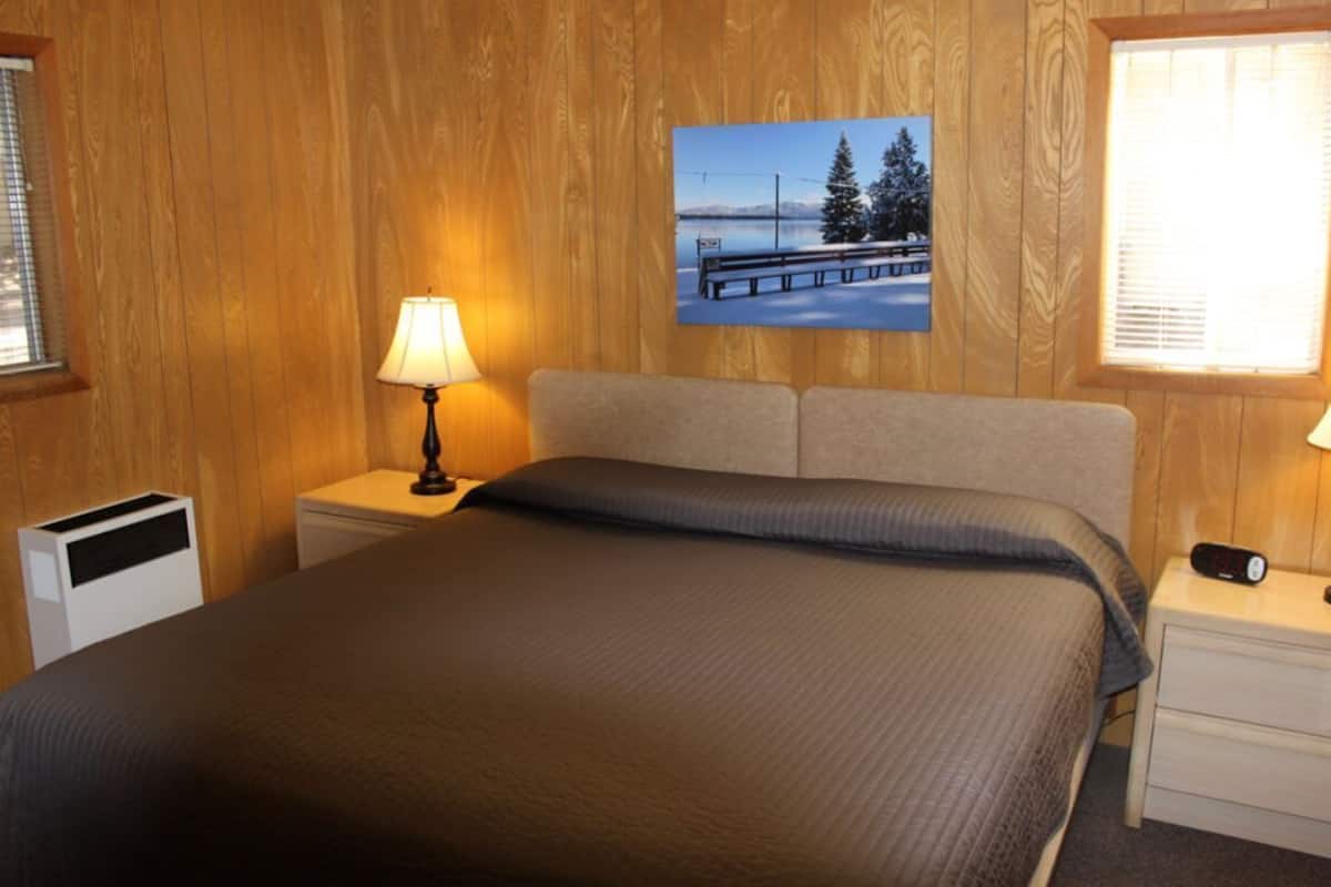 Un dormitorio con una cama, dos mesitas de noche con lámparas, un cuadro en la pared y una ventana con persianas.