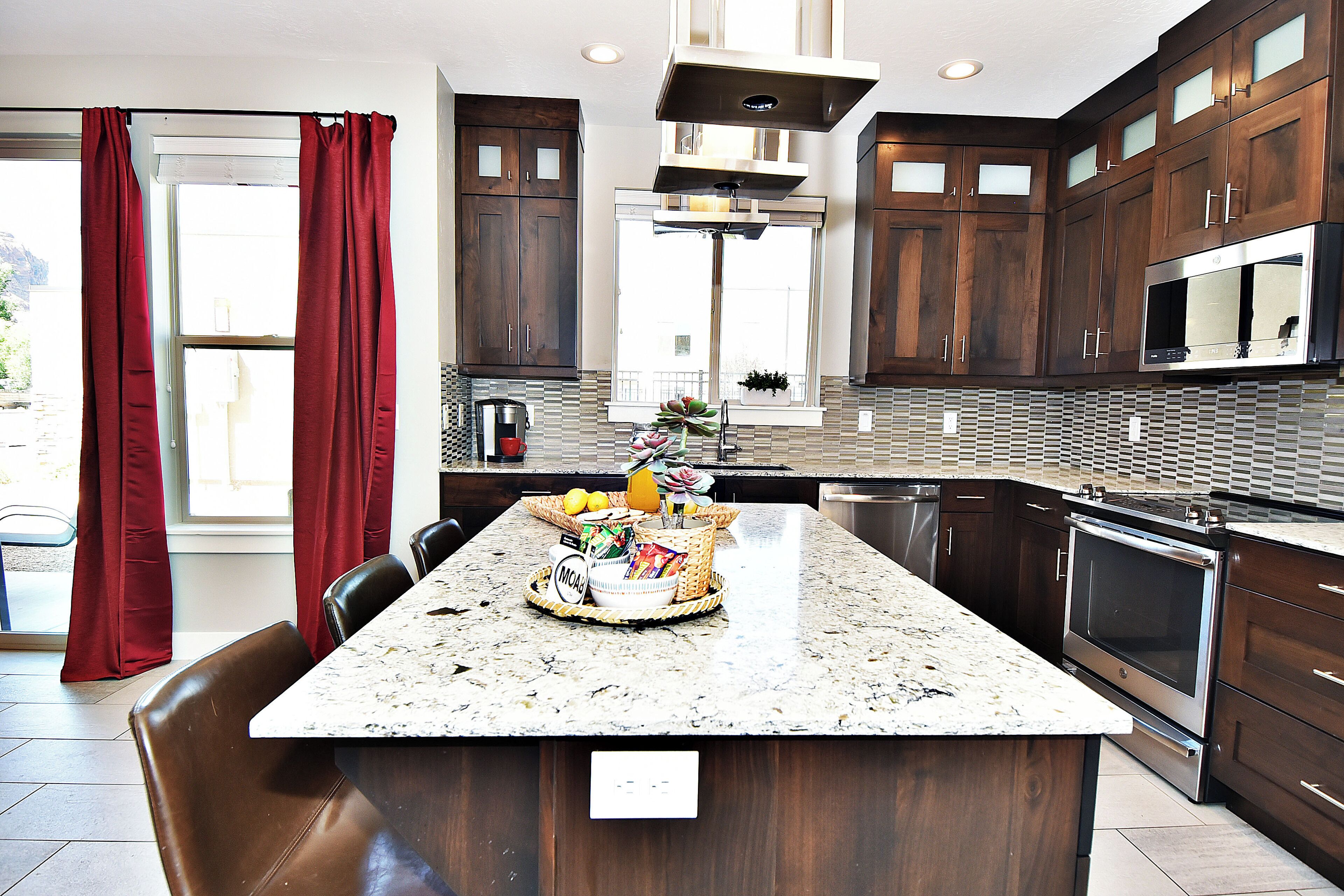 A modern kitchen with dark wood cabinets, a large island with a granite countertop, stainless steel appliances, and a window with red curtains.