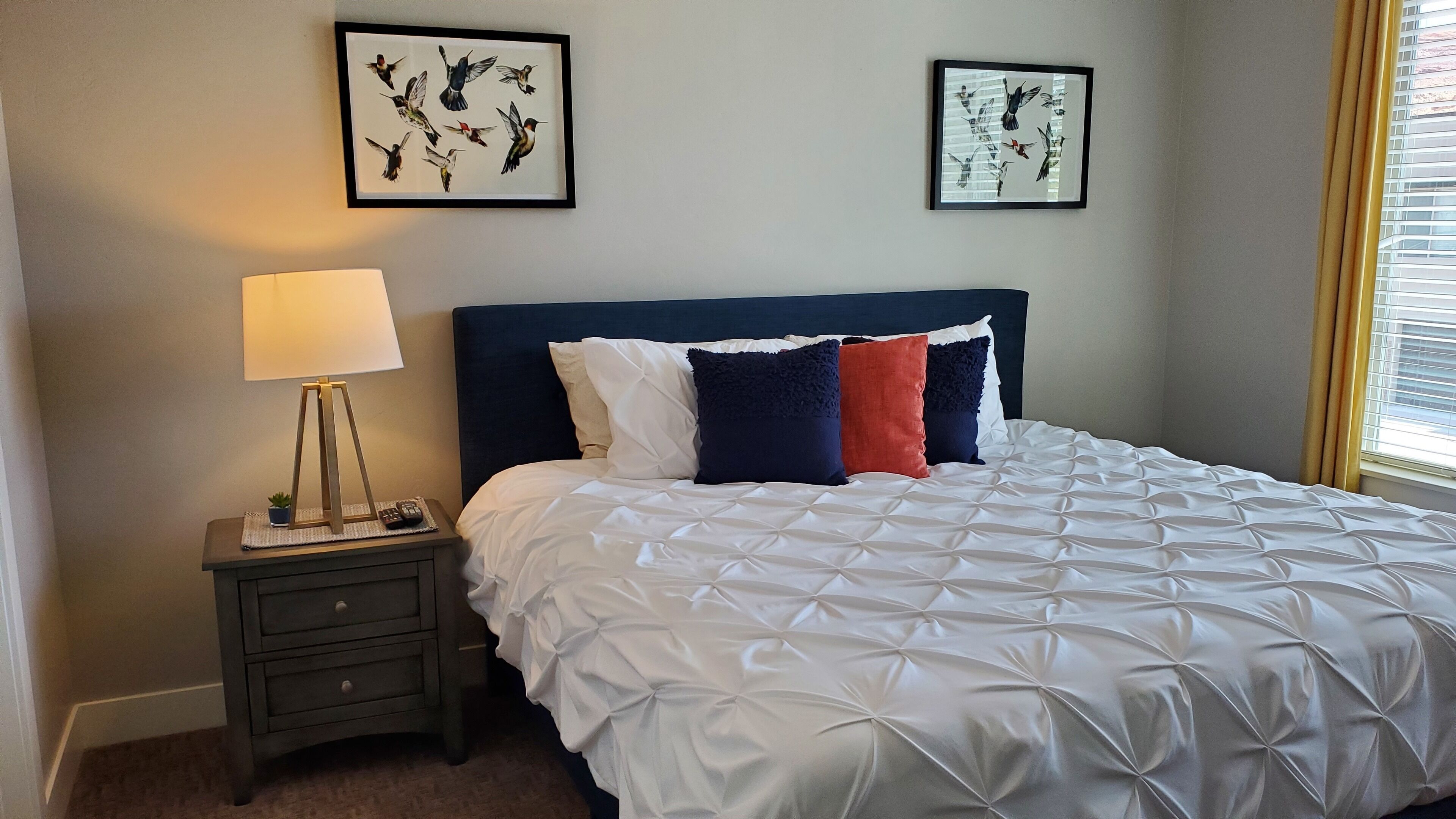 Een net opgemaakt bed met een witte quilt, een donkerblauwe hoofdbord en kleurrijke kussens. Er staat een klein nachtkastje met een lamp, een afstandsbediening en een kleine plant. Twee ingelijste foto’s van vogels hangen aan de muur boven het bed.