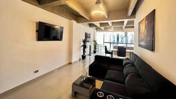 A modern living room with a black sofa, a coffee table, and a flat-screen TV mounted on the wall. There are potted plants and a dining area visible in the background. - The Aragon El Poblado Lofts Triplex w/Penthouse (Medellín)