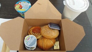 Pahvilaatikossa on erilaisia ruoka-aineita, kuten croissant, voileipä ja purkki jogurttia.