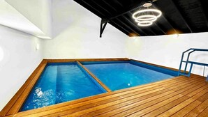 A rectangular indoor swimming pool with wooden deck flooring, surrounded by white walls and a black ceiling with a circular light fixture.