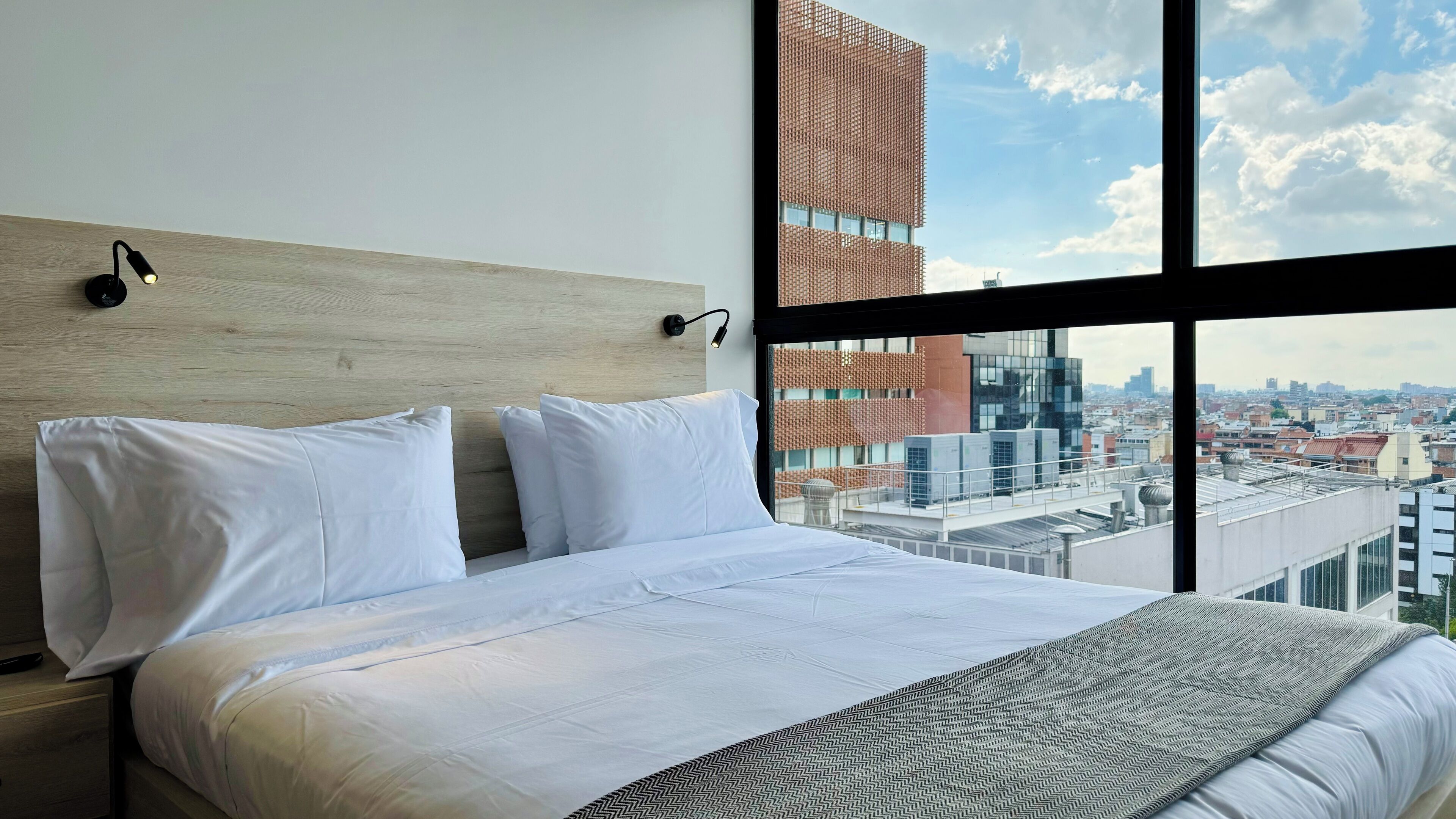 A modern hotel room with a large bed, two bedside lamps, and a city view through a large window.