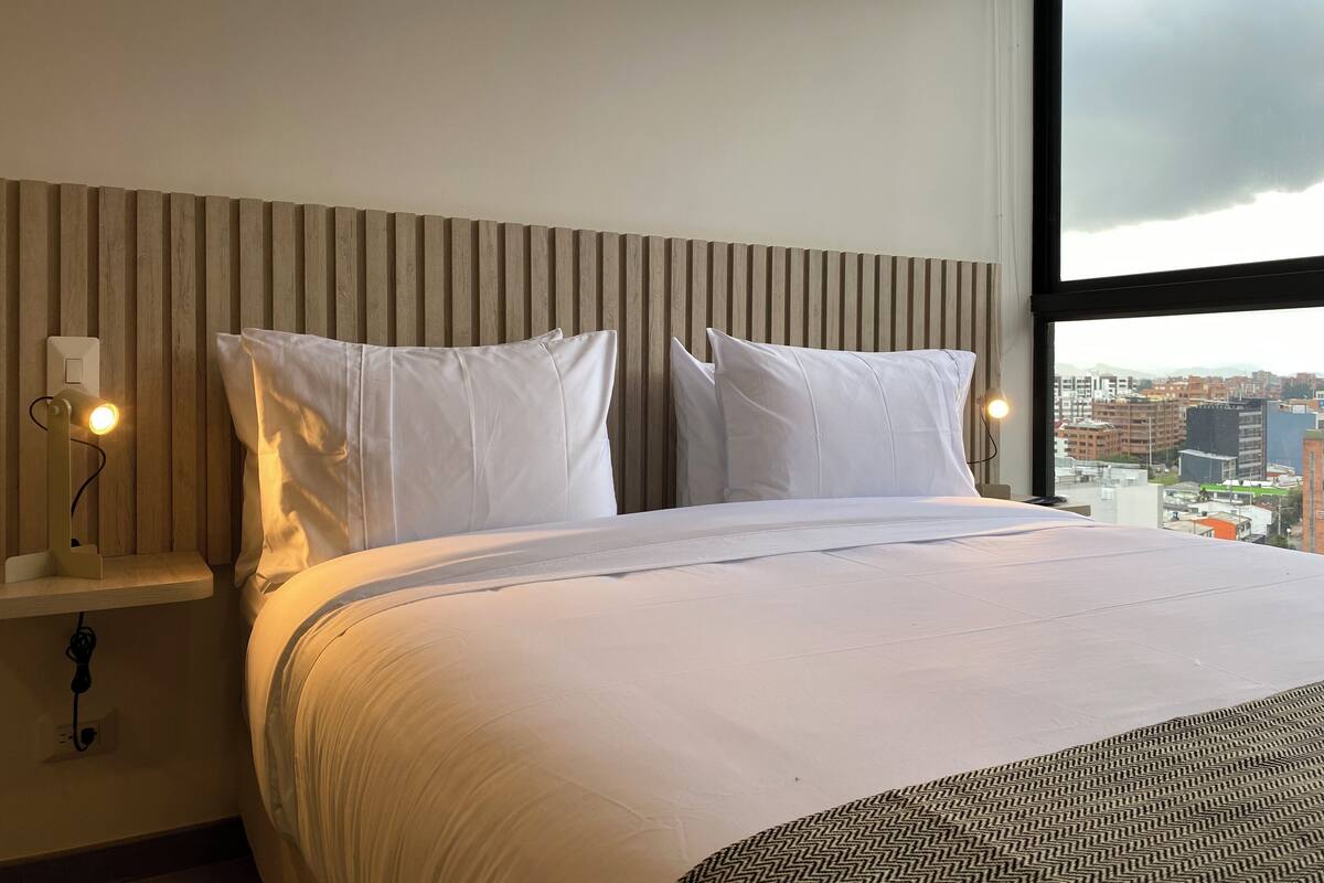 A hotel room with a bed, two white pillows, a wooden headboard, a bedside table with a lamp, and a view of the city through a large window.