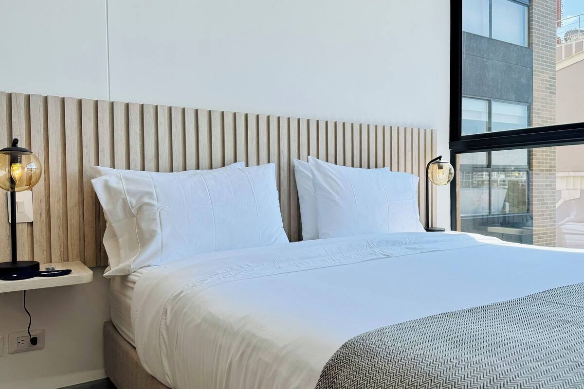 A modern hotel room with a large bed, two bedside lamps, a wooden headboard, and a large window offering a view of the city.