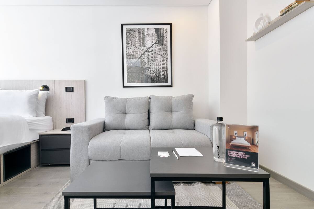 A modern hotel room with a bed, a grey sofa, a small table with a brochure, and a framed cityscape picture on the wall.