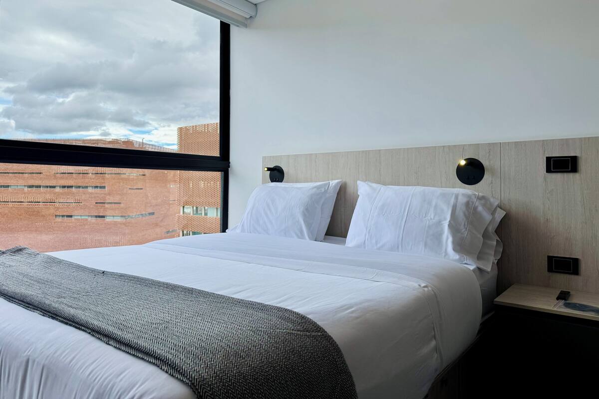 A modern hotel room with a large bed, a wooden headboard, a nightstand, and a view of a brick building through a large window.