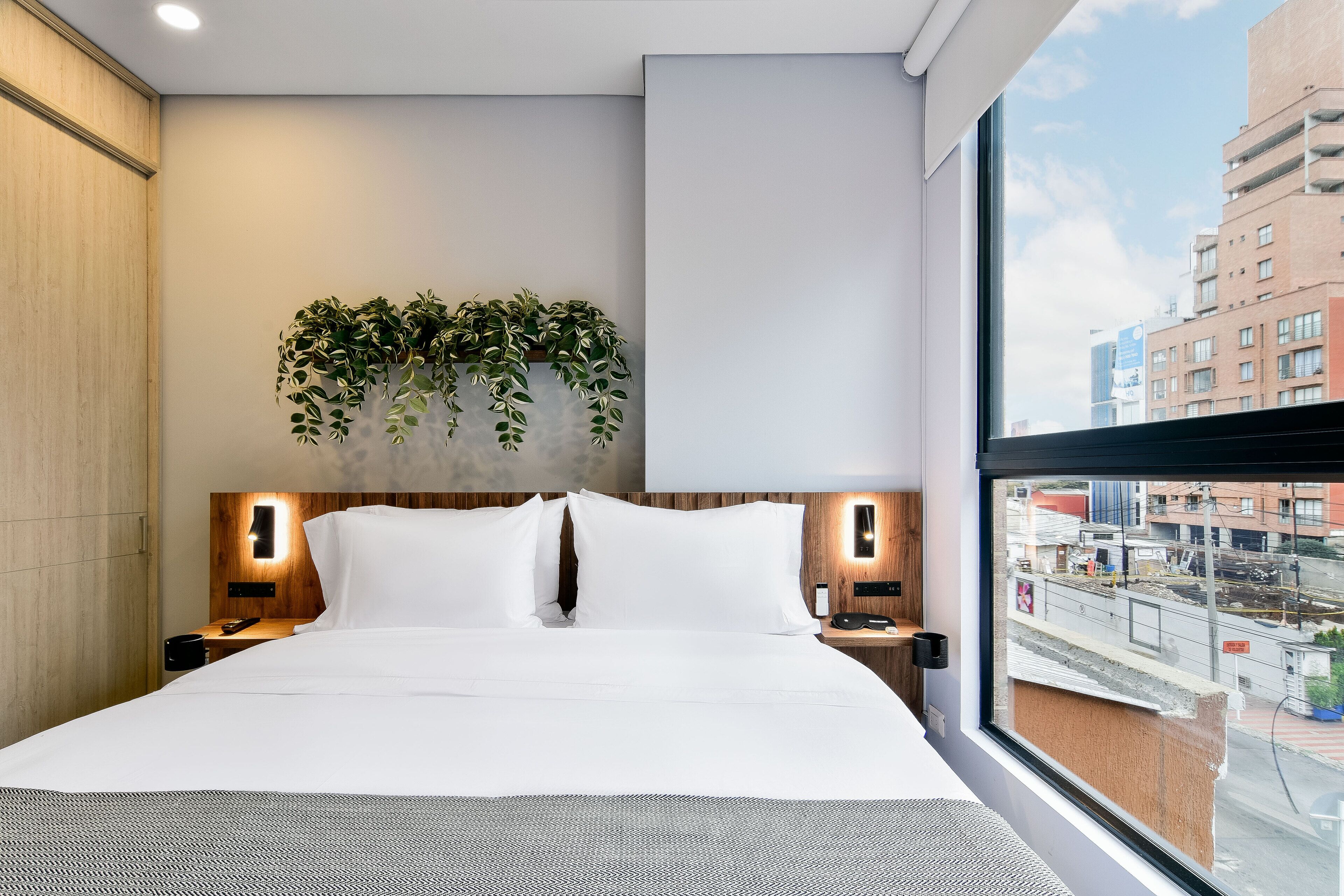 Une chambre d’hôtel moderne avec un grand lit, deux tables de chevet, une plante fixée au mur et une vue sur la ville par une grande fenêtre.