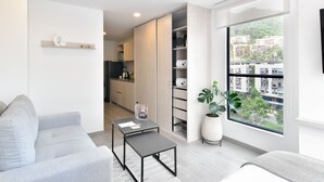 Een moderne woonkamer met een grijze bank, een kleintje tafeltje en een groot raam met uitzicht op gebouwen en groen.