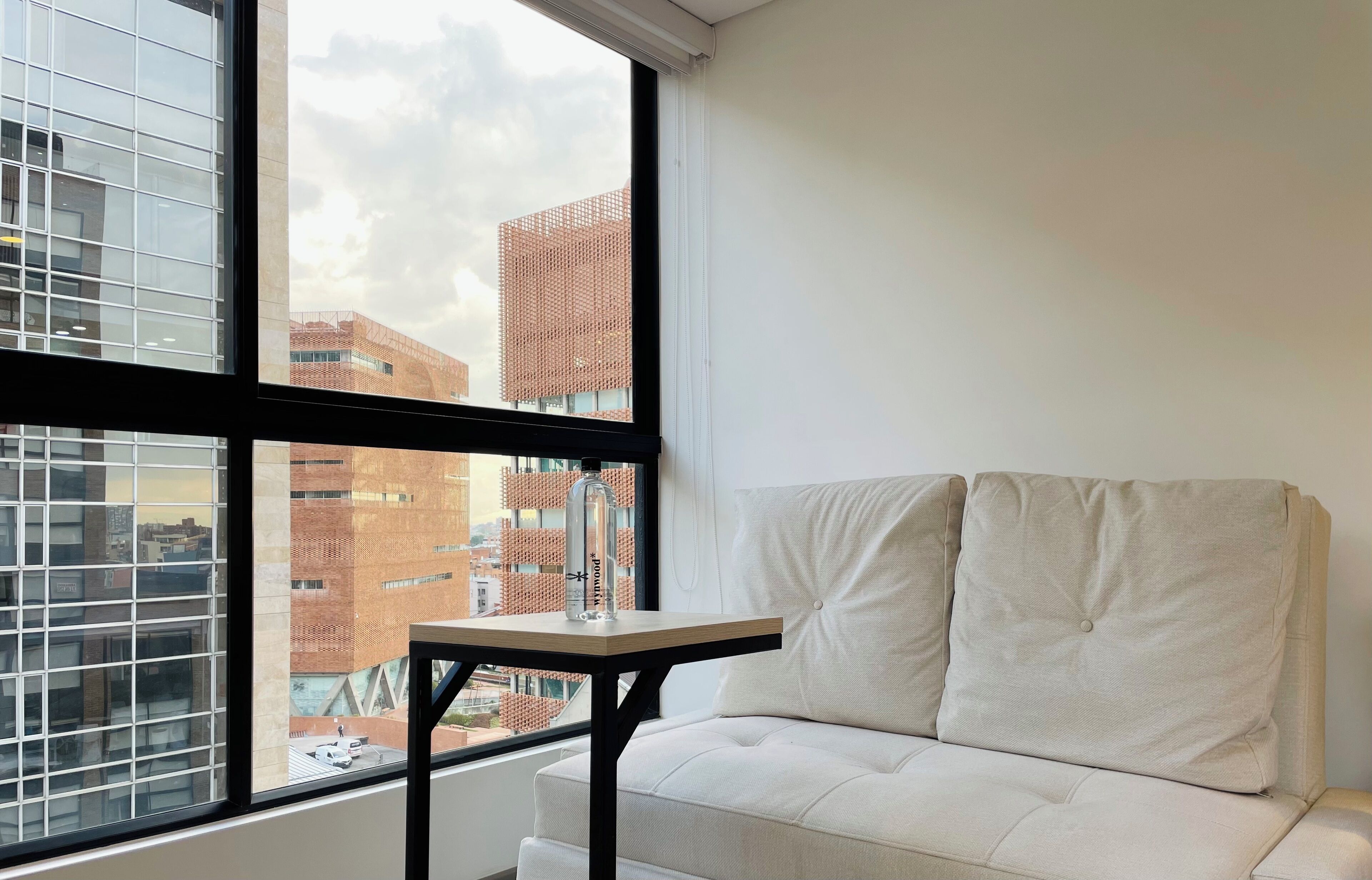 A room with a white sofa and a small table, a large window with a view of city buildings.