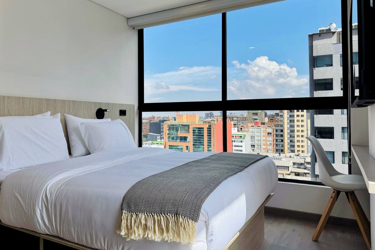 A modern hotel room with a large bed, a view of the cityscape, and a desk with a chair.