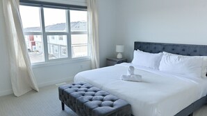 A neatly arranged bedroom with a large bed, a bedside table with a lamp, a window with curtains, and a grey upholstered bench.