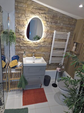 A bathroom with a stone wall, a round mirror, a sink, a towel rack, a plant, and a shower area.