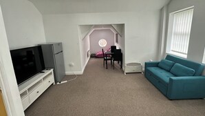 A modern living room with a flat-screen TV, a teal sofa, a dining area with a table and chairs, and a small kitchenette.