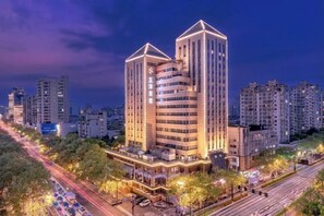 Un hotel moderno con una imponente torre central, rodeado de otros edificios, y una calle concurrida con coches aparcados.