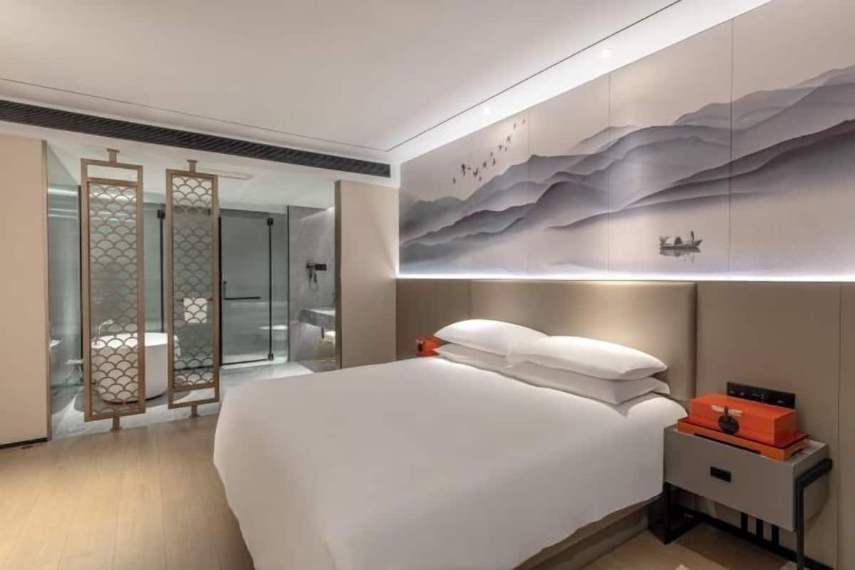 Una habitación de hotel moderna con una cama grande, una mesita de noche, una obra de arte colgada en la pared y una puerta corredera de vidrio que da al baño.