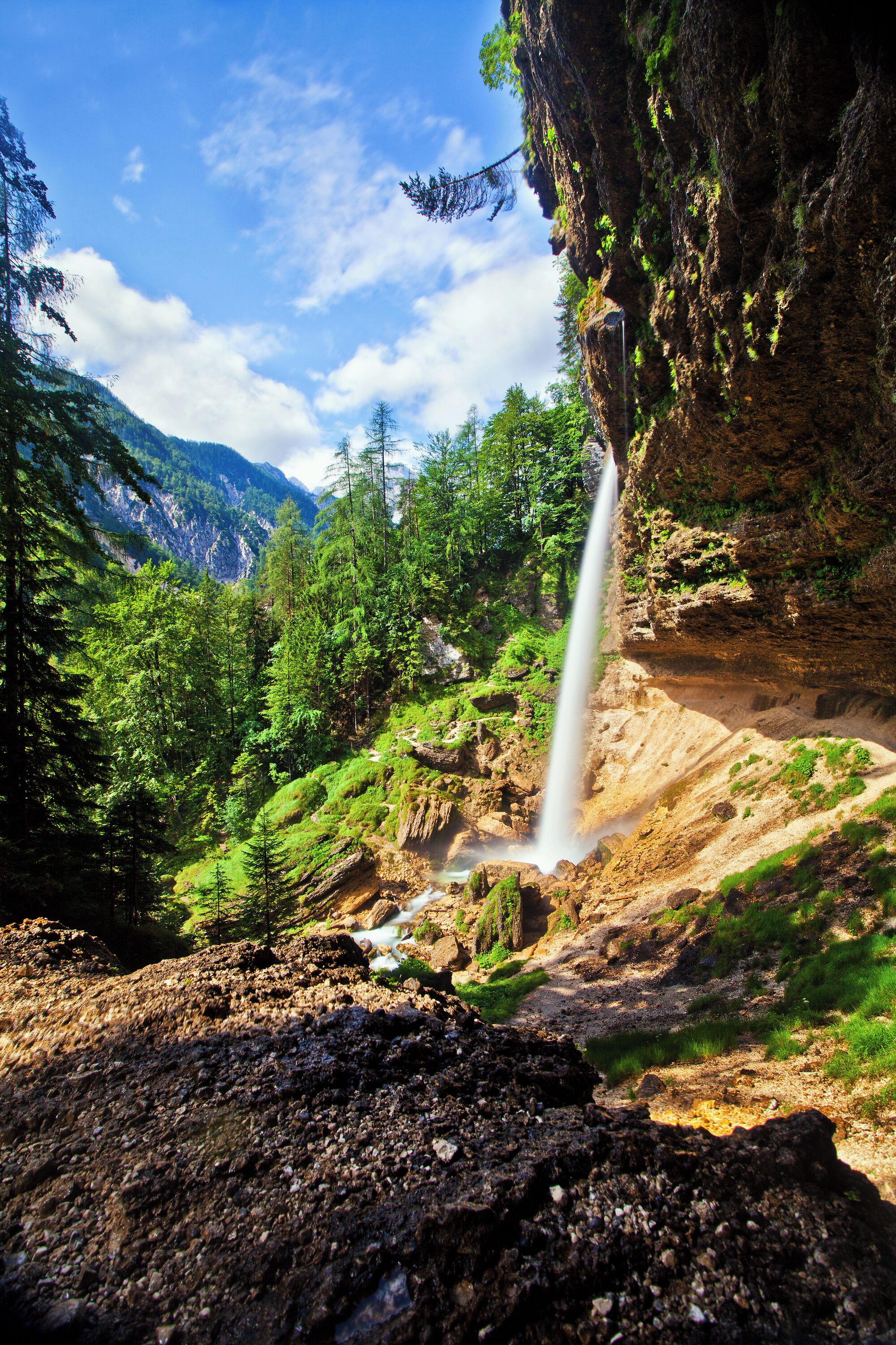 Ein Wasserfall stürzt über eine felsige Klippe, umgeben von üppiger Vegetation und Bergen im Hintergrund.