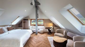 A spacious attic bedroom with a large bed, two armchairs, a wooden coffee table, and a view of the outdoors through skylights.