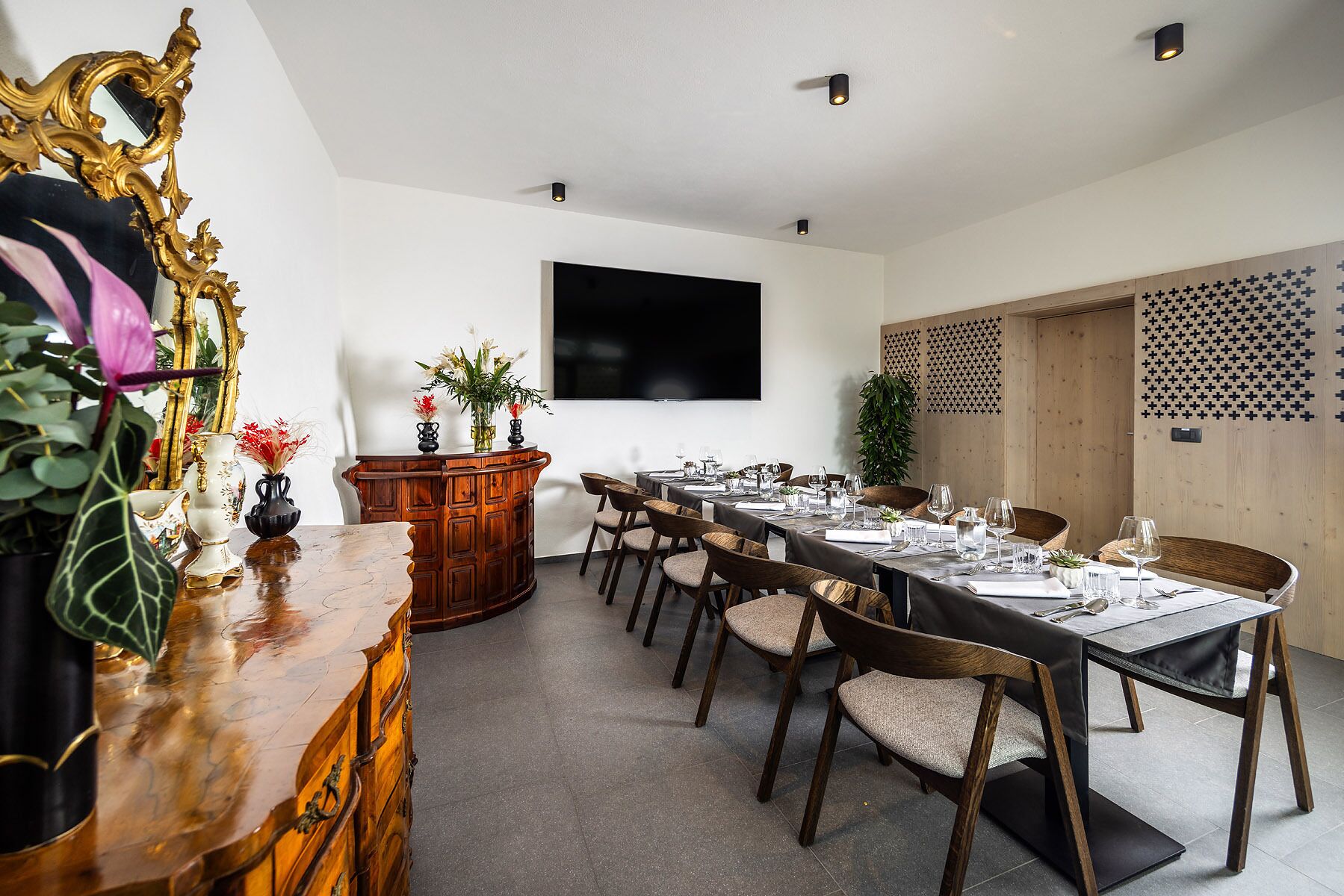 Un comedor con una mesa larga dispuesta para una comida, un televisor montado en la pared y un aparador decorado con plantas.