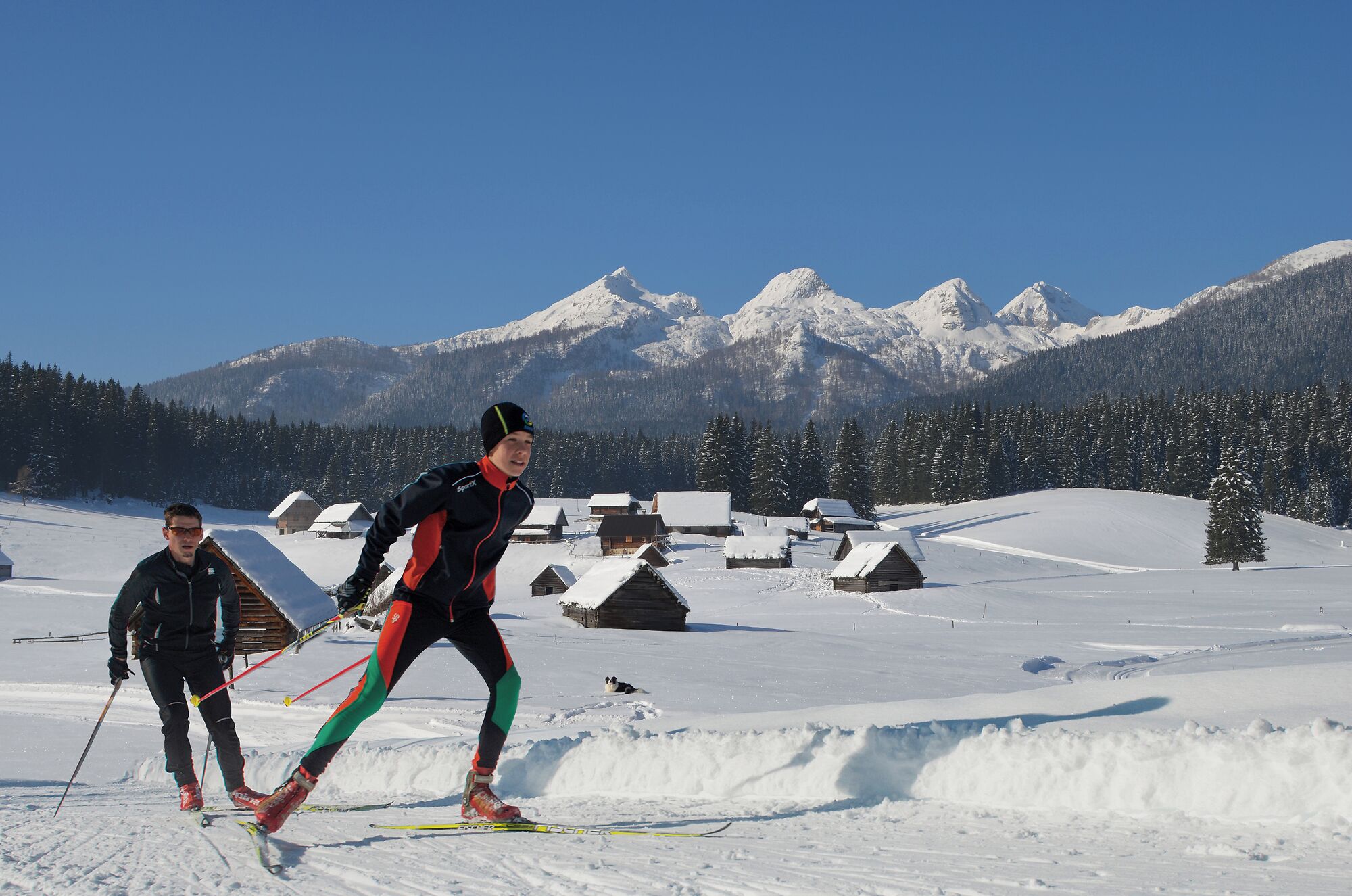 A snowy landscape with a person skiing and another person standing nearby. There are wooden cabins in the background and a mountain range in the distance.