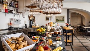 A buffet setup with various food items, including bread, fruits, and cheese, in a well-lit room with a tiled floor and a chandelier.