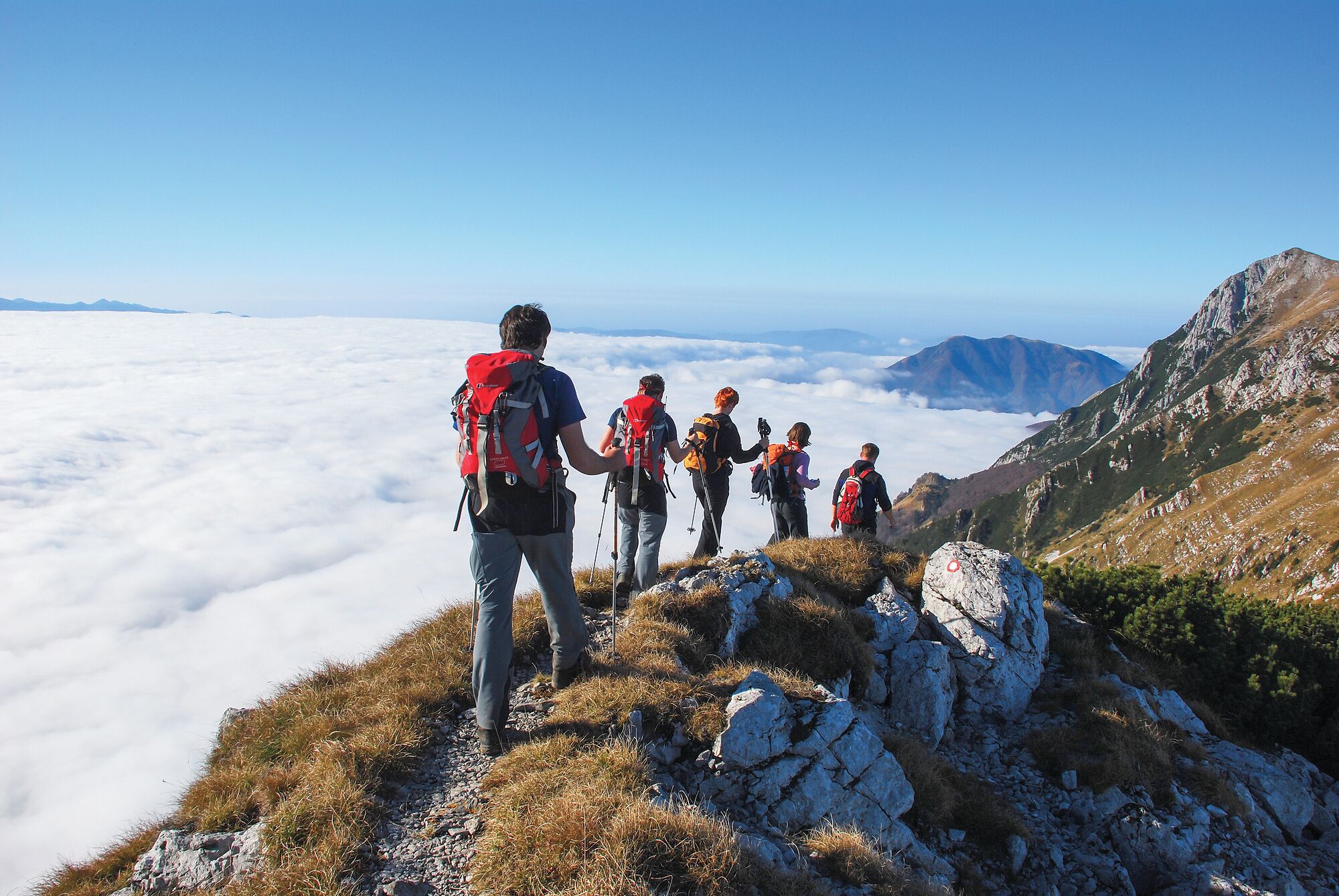 Un grupo de excursionistas en un sendero de montaña con terreno rocoso y una vista de nubes abajo.