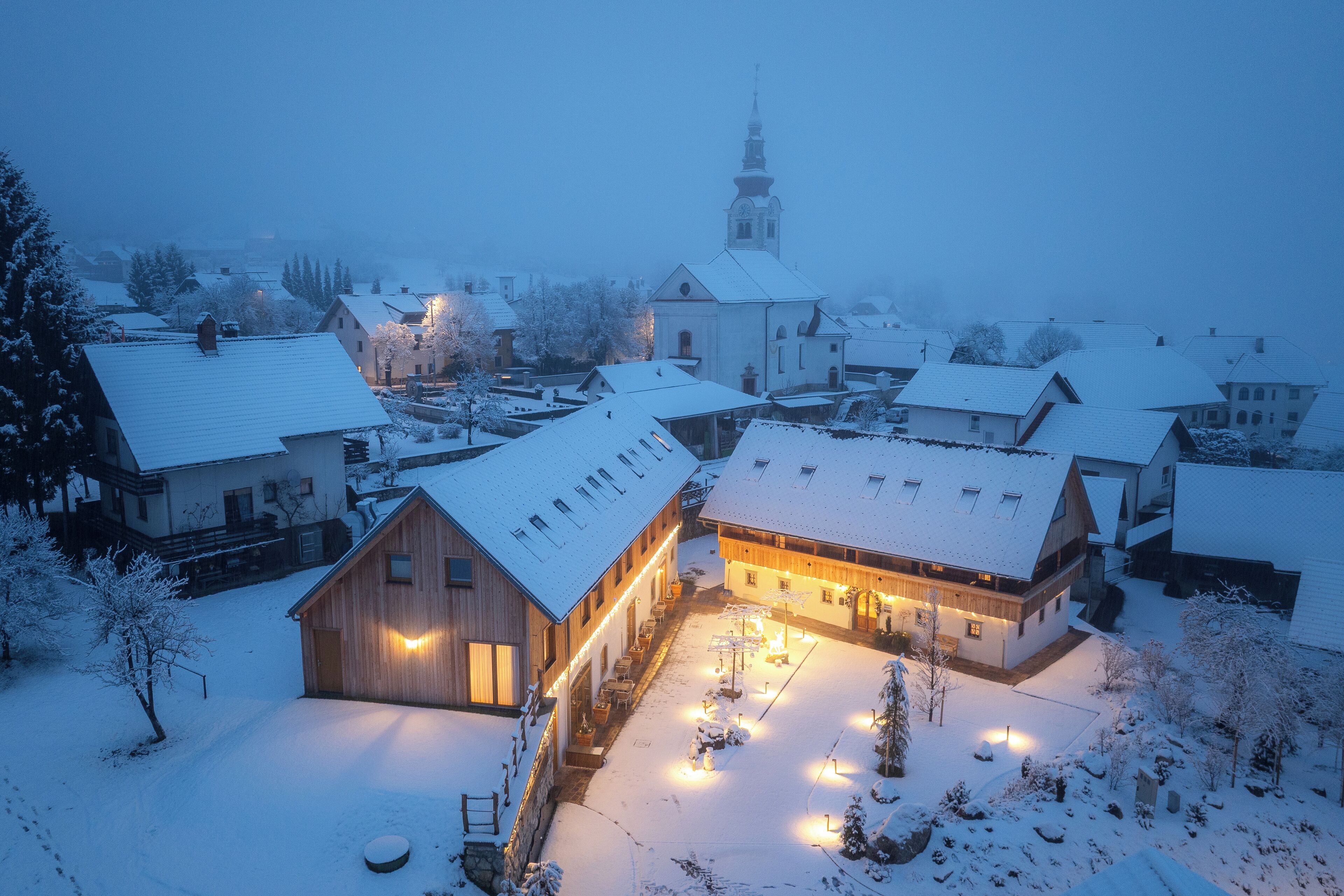 Un pueblo cubierto de nieve con edificios de madera tradicionales, una iglesia con un campanario alto y un edificio central bien iluminado con techo inclinado.