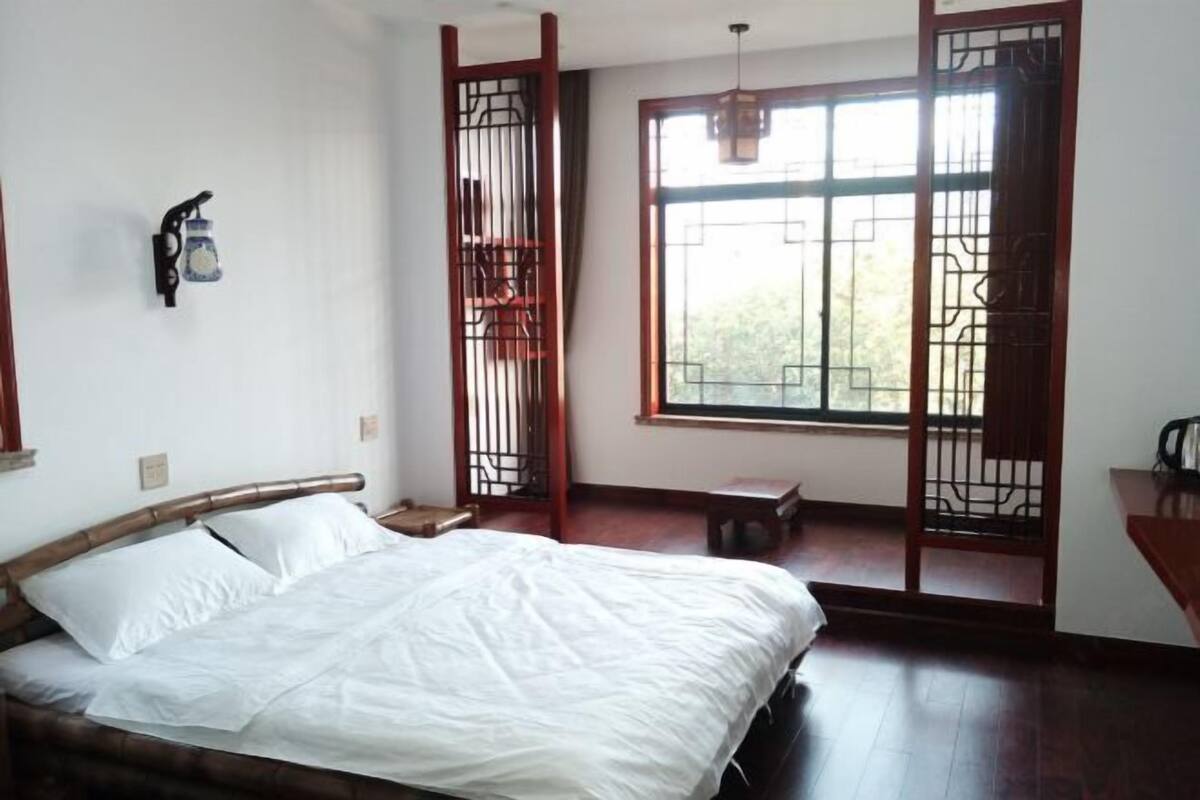 A bedroom with a bed, a window with a view, a nightstand, and a wall-mounted light fixture.