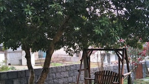 A wooden swing chair under a tree in a garden area with a stone wall and a metal fence in the background. - Moganshan Yashan Inn (Deqing)