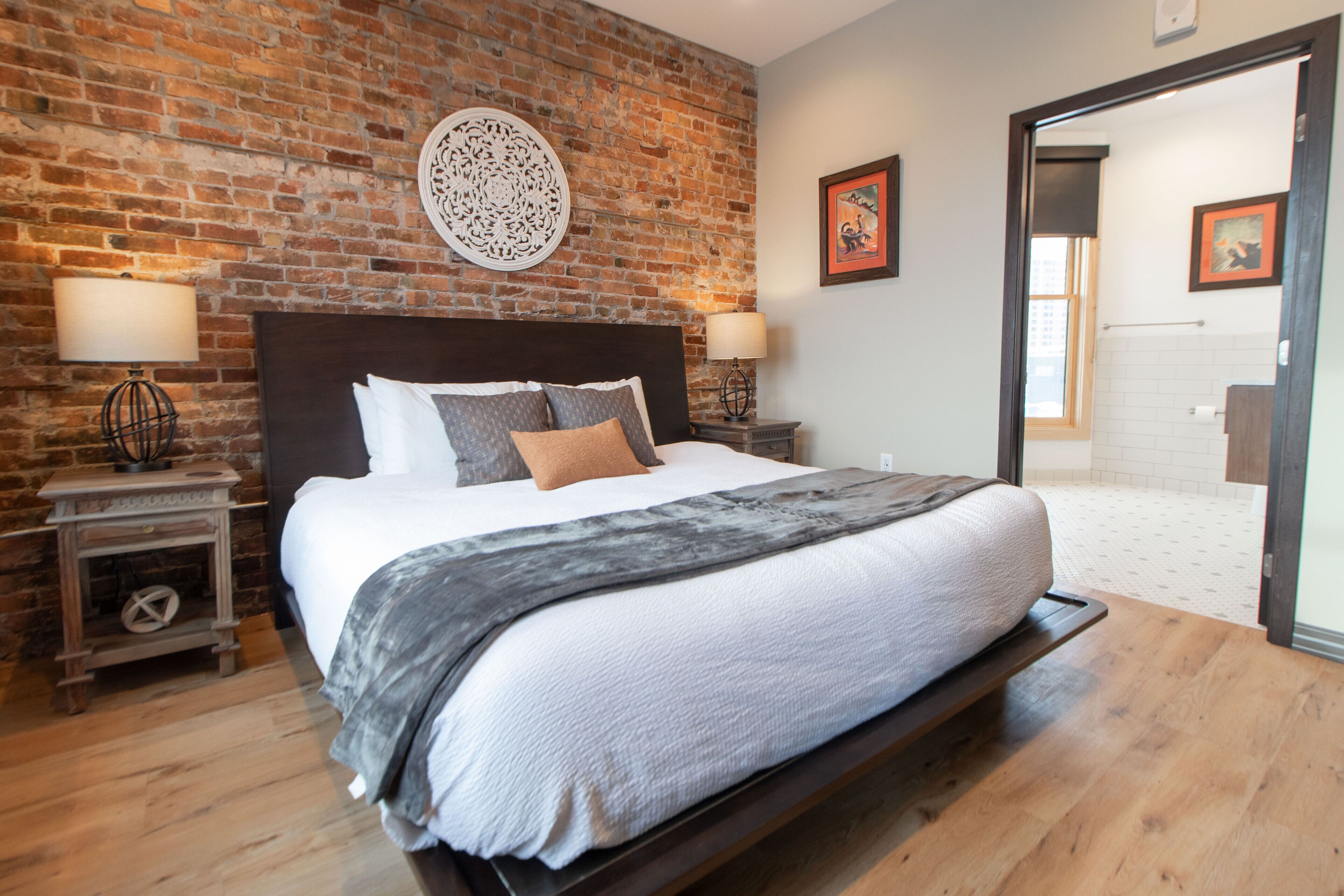 Un dormitorio con pared de ladrillo, una cama grande con cabecera, dos mesitas de noche con lámparas, y una puerta que da al baño.