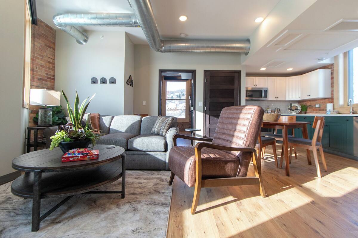 Un salon moderne avec un canapé gris, une chaise en bois et une table basse ronde. On y trouve un mur en briques et une fenêtre donnant sur un paysage.