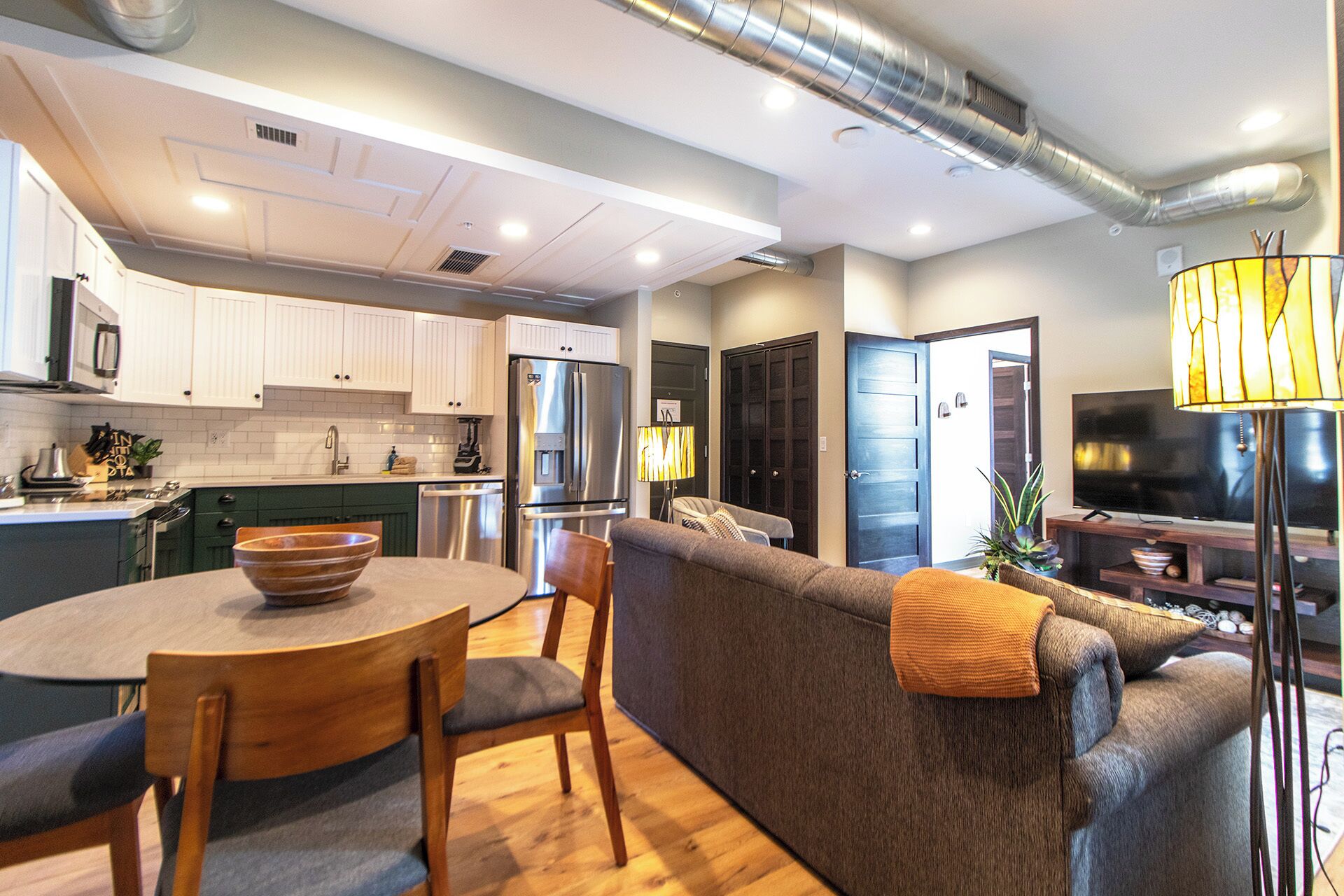 A modern kitchen with white cabinets, a built-in oven, and a stainless steel refrigerator. There is a dining area with a round table and chairs, and a living room with a sofa and a TV.