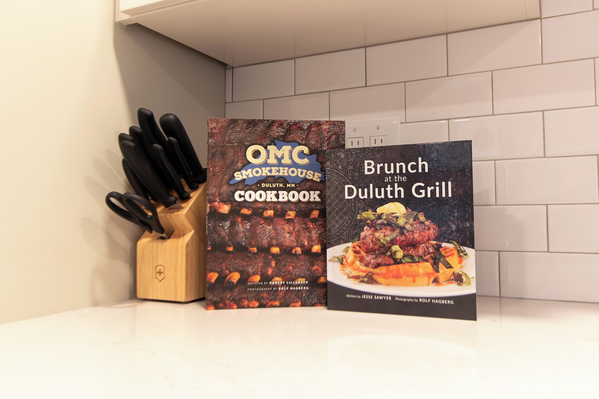Deux livres de recettes sont posés sur un plan de travail de cuisine, à côté d’un bloc à couteaux : l’un s’intitule « OMC Smokehouse Duluth, MN Cookbook » et l’autre « Brunch at the Duluth Grill ».