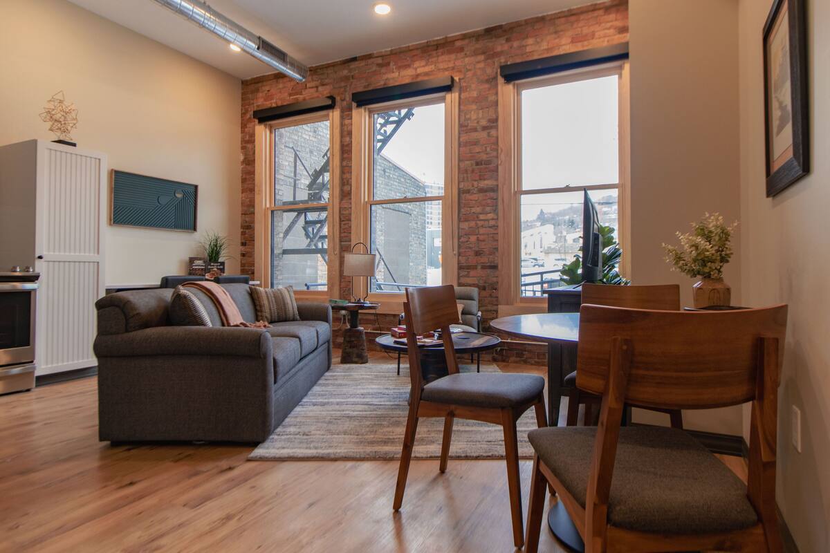 Un salon moderne avec un mur en briques, de grandes fenêtres, un coin repas et une cuisine.