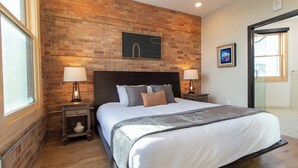 Une chambre avec un mur en briques, un grand lit, deux tables de chevet avec des lampes et un tableau au mur.