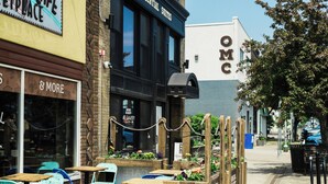 Espace extérieur aménagé avec des tables et des chaises, un bâtiment portant une enseigne avec l’inscription « OMC », et une devanture de magasin avec une enseigne indiquant « RAFRAÎCHISSEMENTS ».