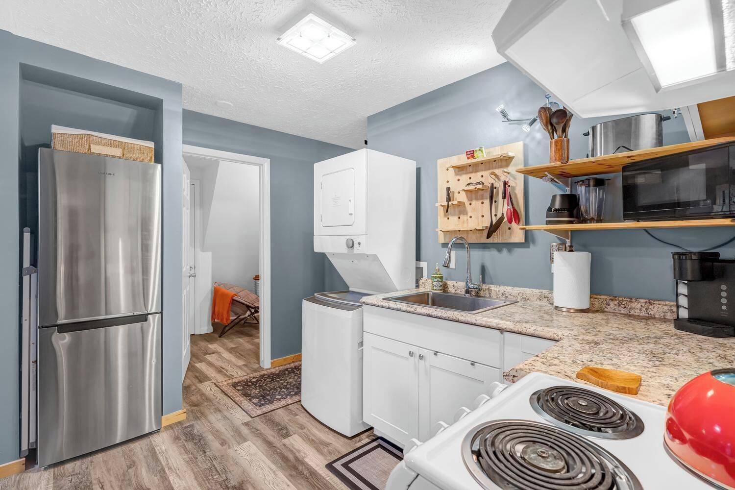 A compact kitchen with a stainless steel refrigerator, white cabinets, a microwave, and a red kettle.