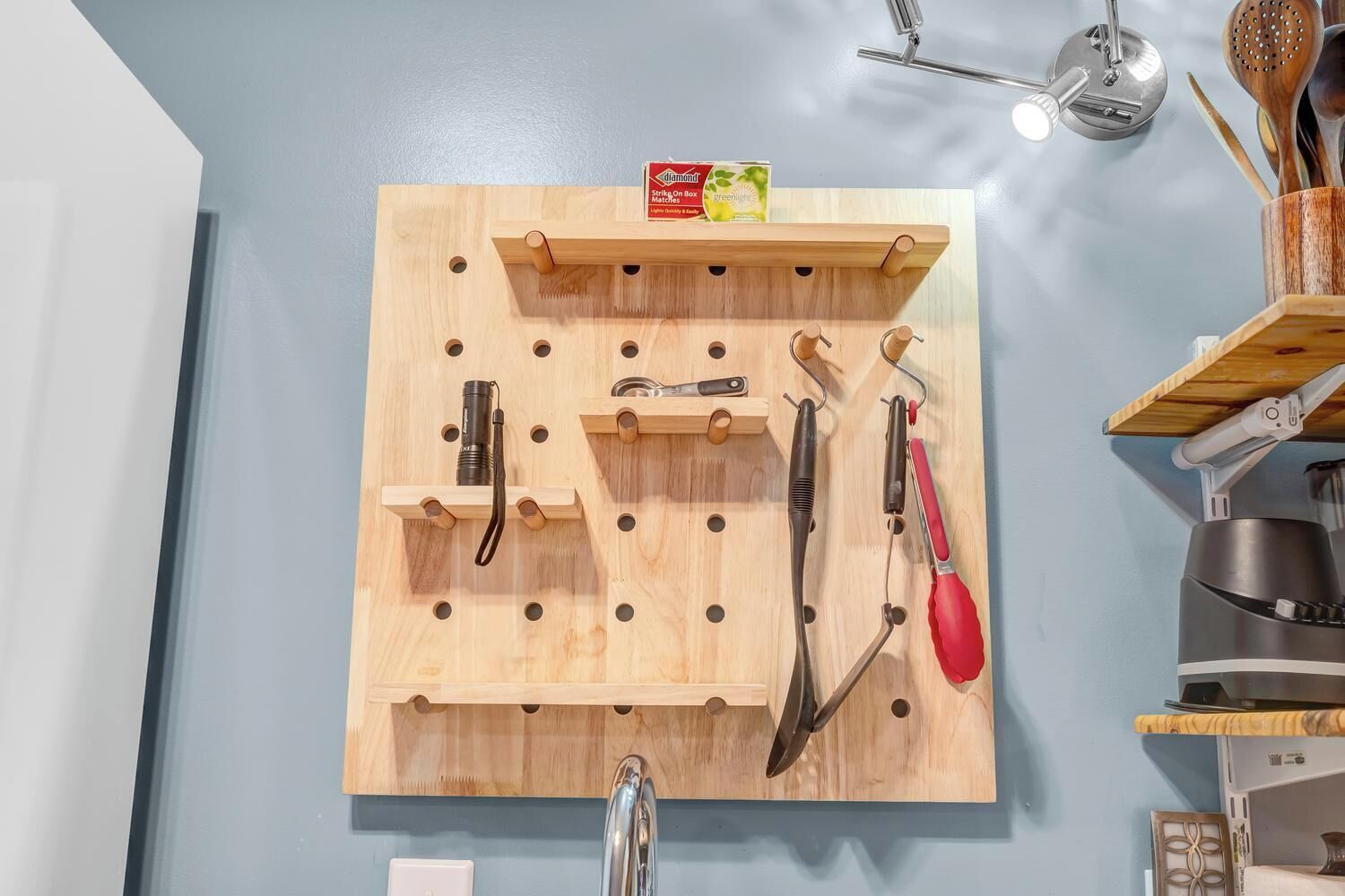 A kitchen wall-mounted pegboard with various utensils and a box of Diamond Strike Match on Box.