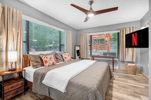 A hotel room with a bed, bedside table, lamp, TV, and large windows with blinds.