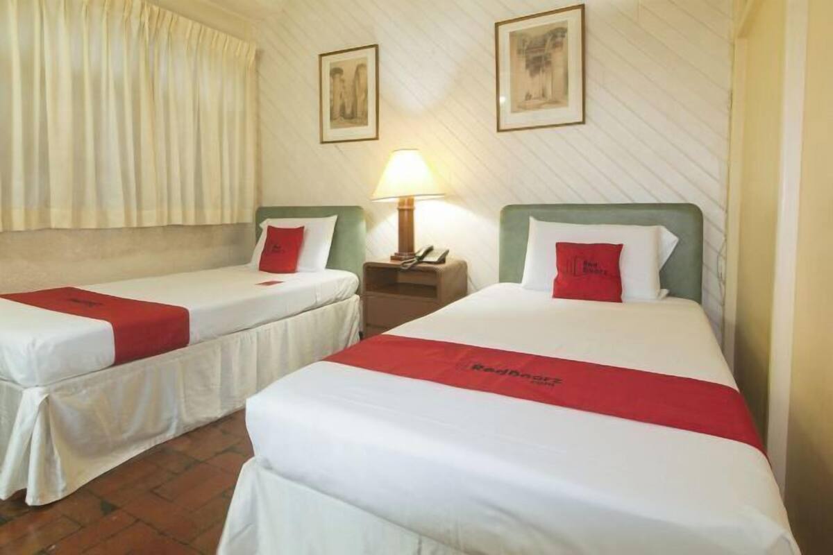 Habitación de hotel con dos camas, cada una con colcha roja y ropa de cama blanca. Entre las camas, una mesita de noche con una lámpara.