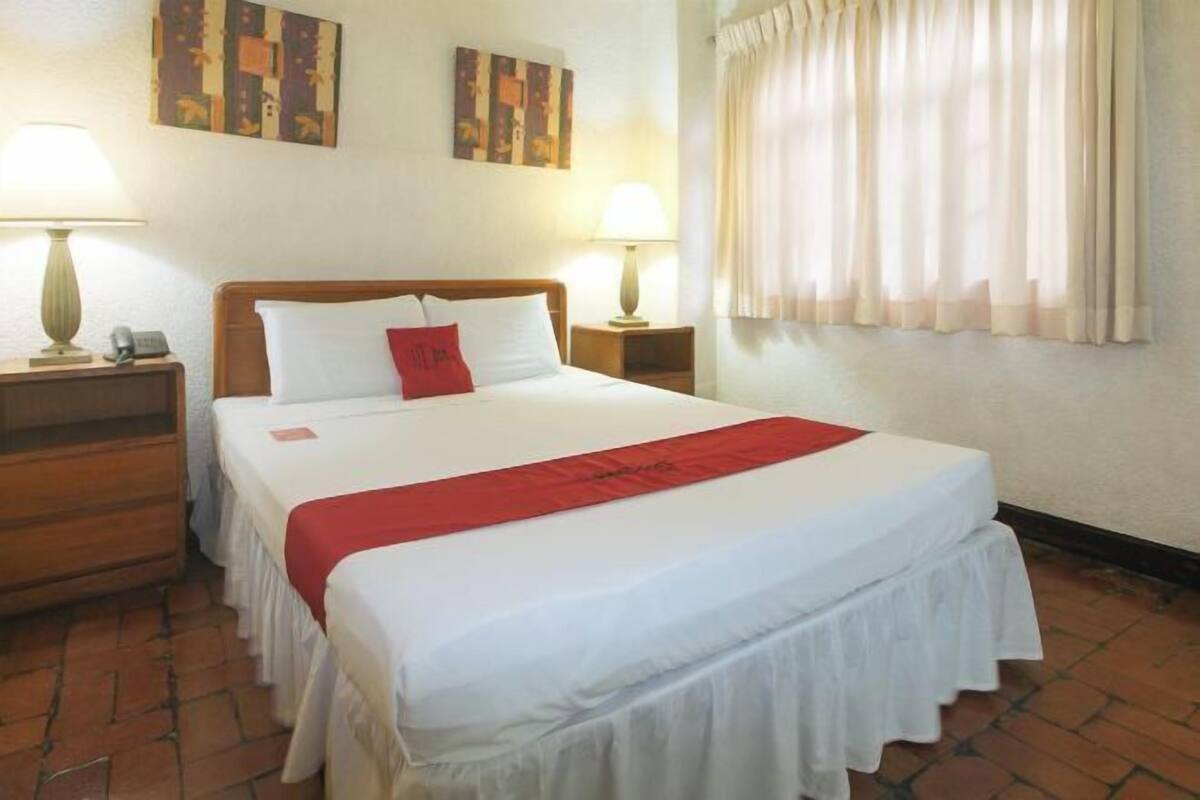 Habitación de hotel con una cama doble, dos mesitas de noche con lámparas, un plaid rojo y obras de arte en las paredes.