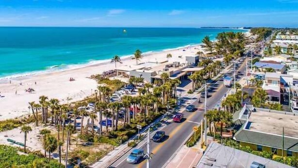 Una calle costera con acceso a la playa, palmeras y coches estacionados.