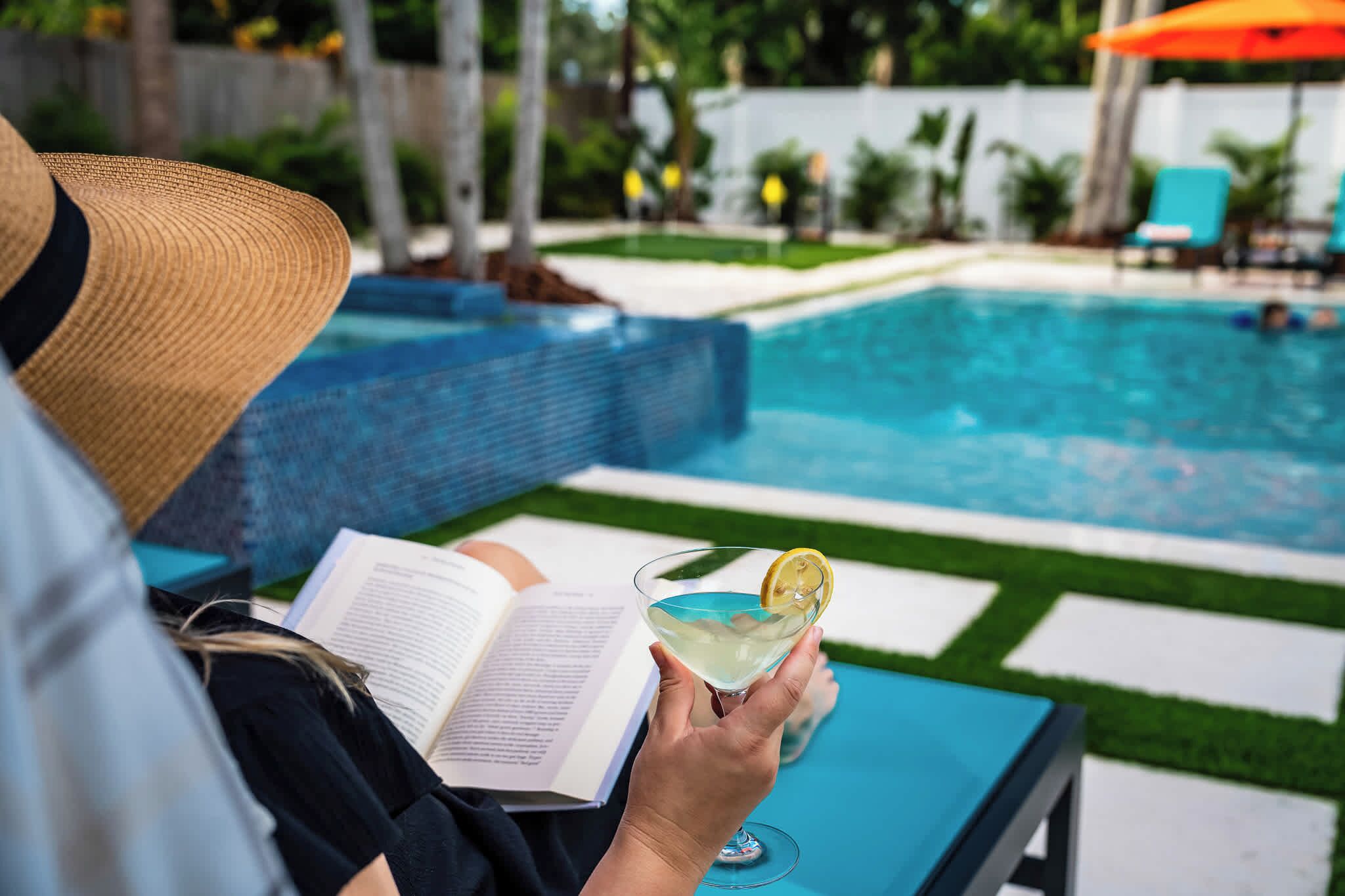 A person reading a book by a pool with a drink in hand.