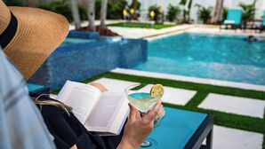Una persona leyendo un libro junto a una piscina, sosteniendo una bebida.