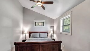 A bedroom with a wooden bed, two bedside lamps, a ceiling fan, a window with blinds, and a small window above the bed.