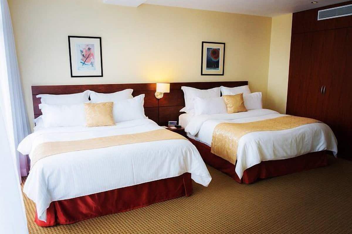 Habitación de hotel con dos camas, cada una con ropa blanca y falda roja. Hay dos lámparas de pared y dos cuadros enmarcados.