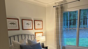 Un dormitorio con cama, ventilador de techo, cuadros de peces enmarcados, mesita de noche con lámpara y ventana con cortinas.